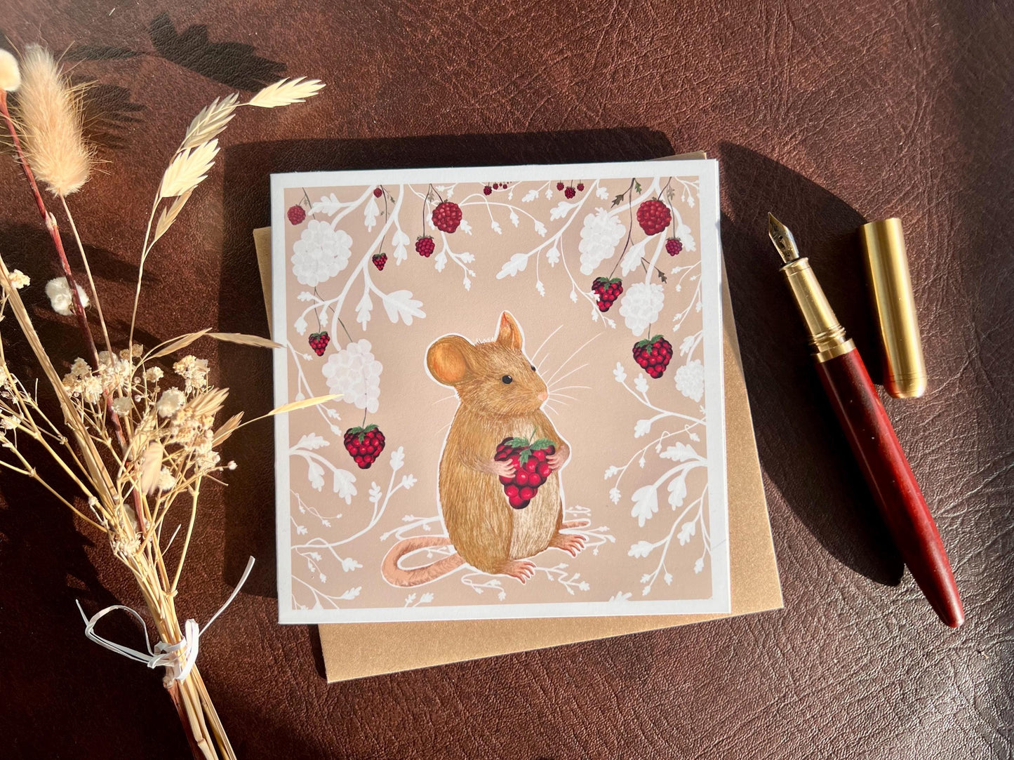 Flat lay image of an artisan greeting card featuring a mouse holding a blackberry surrounded by a white bramble bush pattern with colourful blackberries on a pale pink background, shown laid flat next to a fountain pen and dried flowers in a styled composition.