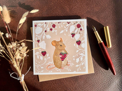 Flat lay image of an artisan greeting card featuring a mouse holding a blackberry surrounded by a white bramble bush pattern with colourful blackberries on a pale pink background, shown laid flat next to a fountain pen and dried flowers in a styled composition.
