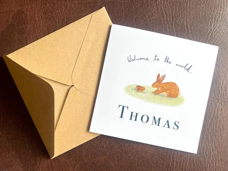 Artisan new baby card featuring a storybook style rabbit illustration shown laid flat on a leatherette tabletop with its kraft envelope, presented in a clean flat lay composition.