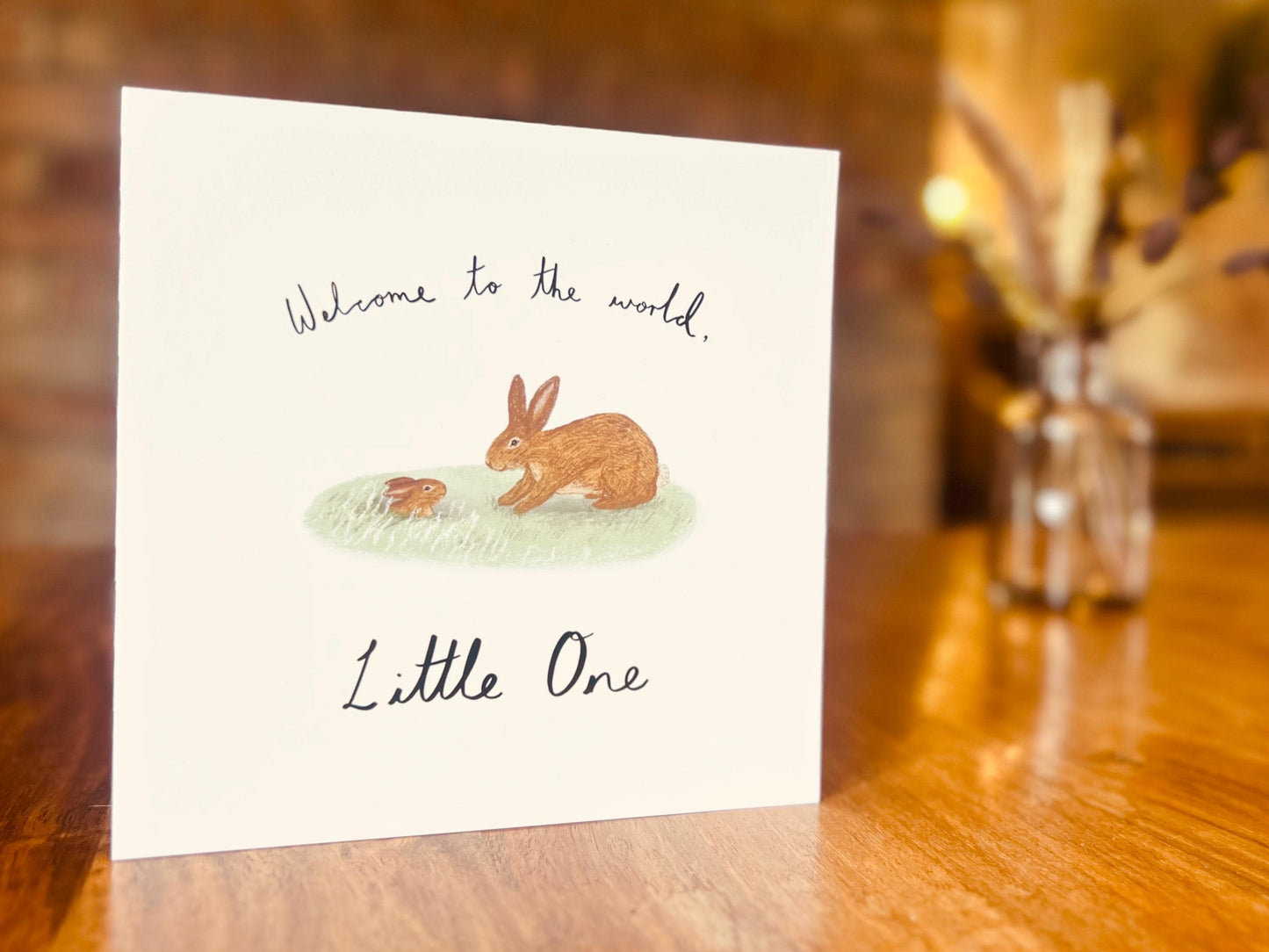 Artisan new baby card displayed upright on a tabletop, featuring a storybook style illustration of a brown rabbit welcoming a baby bunny emerging from a burrow. The card reads 'Welcome to the world, Little One' on the front in a clean lifestyle product shot.