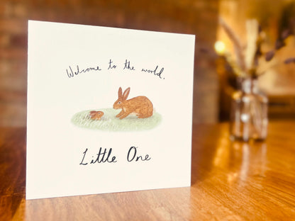 Artisan new baby card displayed upright on a tabletop, featuring a storybook style illustration of a brown rabbit welcoming a baby bunny emerging from a burrow. The card reads 'Welcome to the world, Little One' on the front in a clean lifestyle product shot.