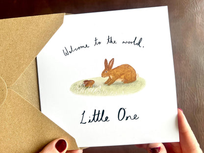 Hands holding an artisan new baby card featuring a storybook style illustration of a rabbit welcoming a baby bunny emerging from a burrow. The card reads 'Welcome to the world, Little One' and is shown as a congratulatory message for new parents.