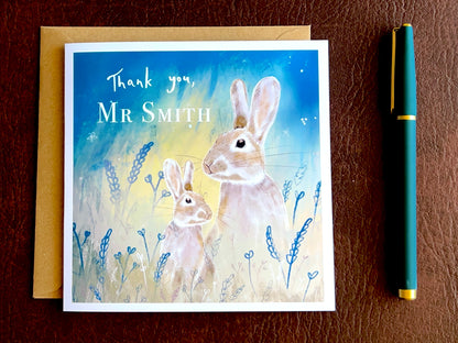 Flat lay image of an artisan thank you teacher card featuring an adult rabbit and young rabbit under a starry blue and green sky, shown laid flat on a tabletop with a kraft envelope. The card reads 'Thank you' followed by a custom teacher name for personalisation.