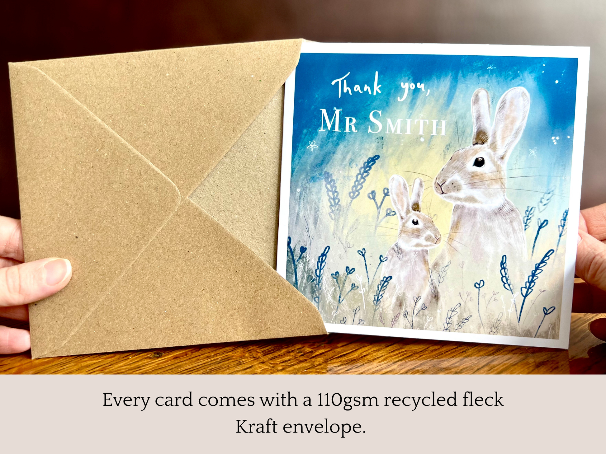 Hands holding an artisan thank you teacher card being removed from a kraft envelope, featuring an adult rabbit and young rabbit under a starry blue and green sky. The card shows 'Thank you' followed by a custom teacher name, with text underneath explaining that every card comes with a 110gsm recycled fleck kraft envelope.
