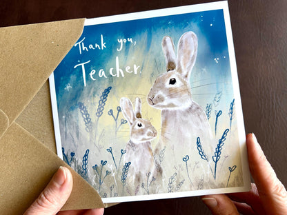 Hands holding an artisan thank you teacher card being removed from a kraft envelope, featuring an illustration of an adult rabbit and young rabbit under a starry blue and green sky. The card reads 'Thank you, Teacher' in a personalised appreciation design.