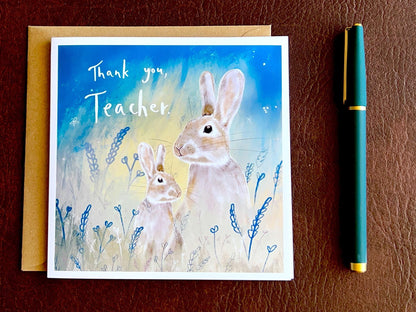 Flat lay image of an artisan thank you teacher card featuring an adult rabbit and young rabbit under a starry blue and green sky, shown laid flat on a tabletop with a kraft envelope. The card reads 'Thank you, Teacher' as a non-personalised version.