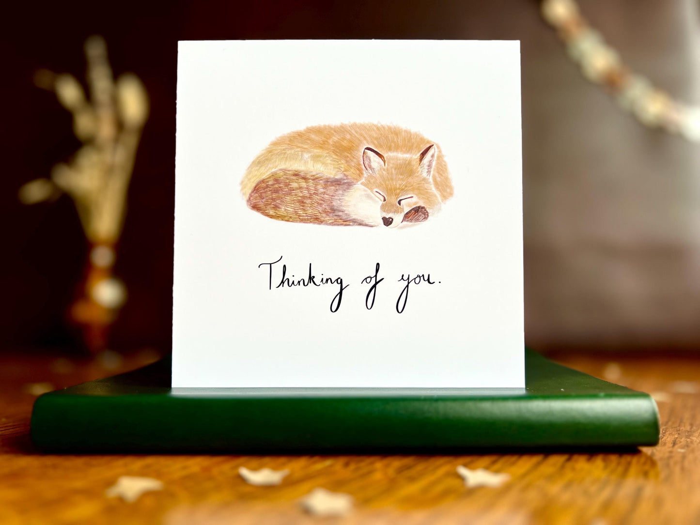 Artisan sympathy card featuring a sleeping fox illustration on a plain white background with handwritten style text reading 'Thinking of you', shown standing upright on a green leatherette book in a styled lifestyle display.