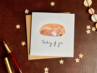 Flat lay image of an artisan sympathy card featuring a sleeping fox illustration on a plain white background with handwritten style text reading 'Thinking of you', shown laid flat on top of a kraft envelope in a simple styled composition.
