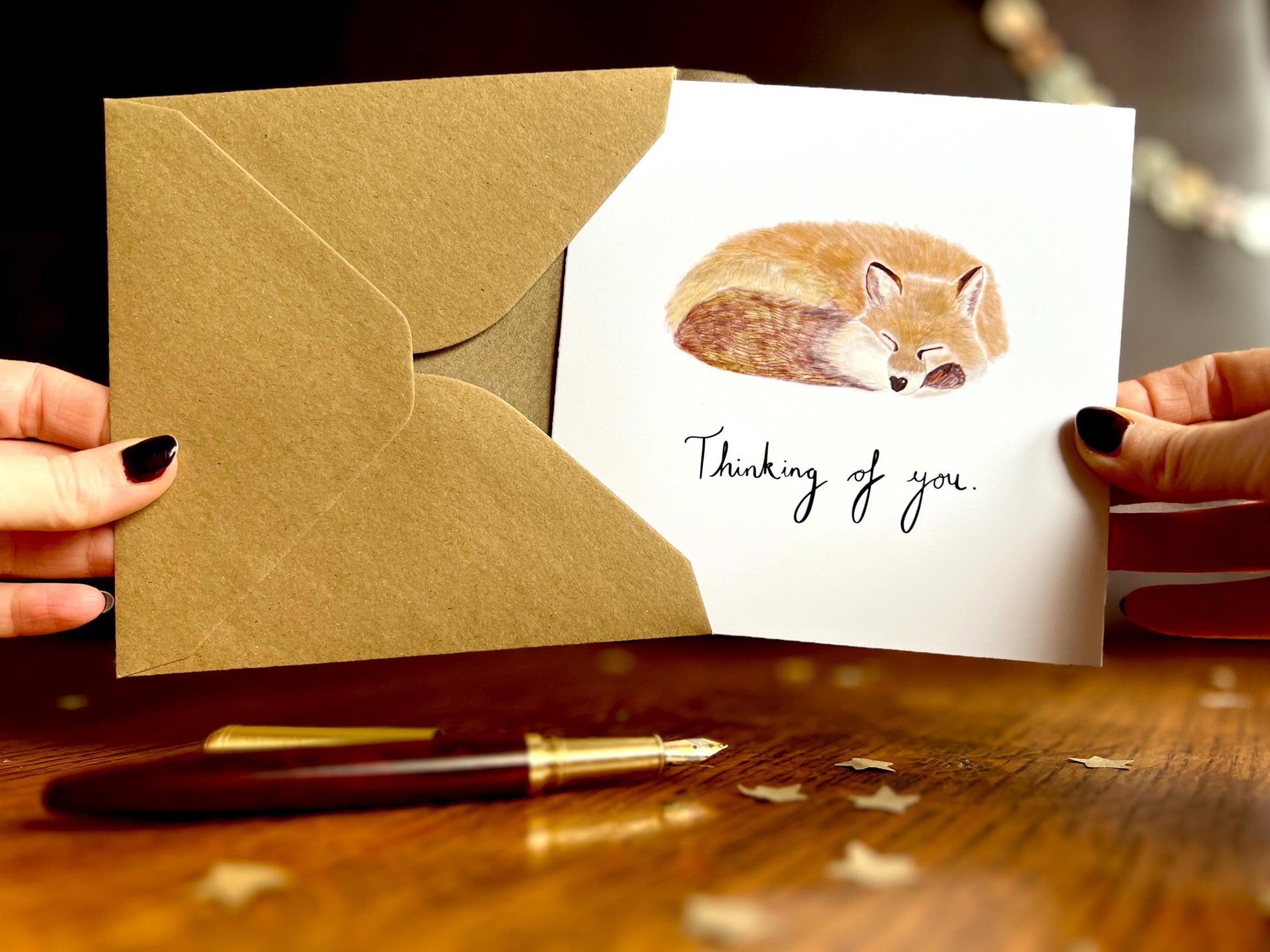 Front view of hands holding an artisan sympathy card being removed from a kraft envelope, featuring a sleeping fox illustration on a plain white background with handwritten style text reading 'Thinking of you'.
