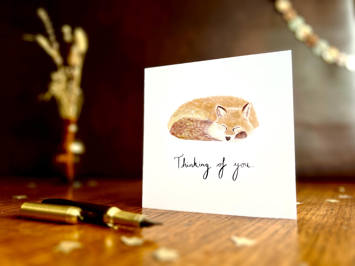 Artisan sympathy card featuring a sleeping fox illustration on a plain white background with handwritten style text reading 'Thinking of you', shown standing upright on a tabletop in a simple lifestyle display.