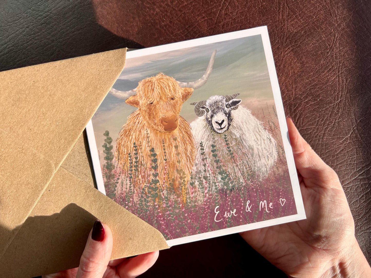 Hands holding an artisan Valentines or anniversary card being removed from a kraft envelope, featuring a playful illustration of a highland cow and Swaledale sheep standing among purple heather with a sage green moorland background. The card includes handwritten style text reading 'Ewe & Me' celebrating love, friendship and companionship.