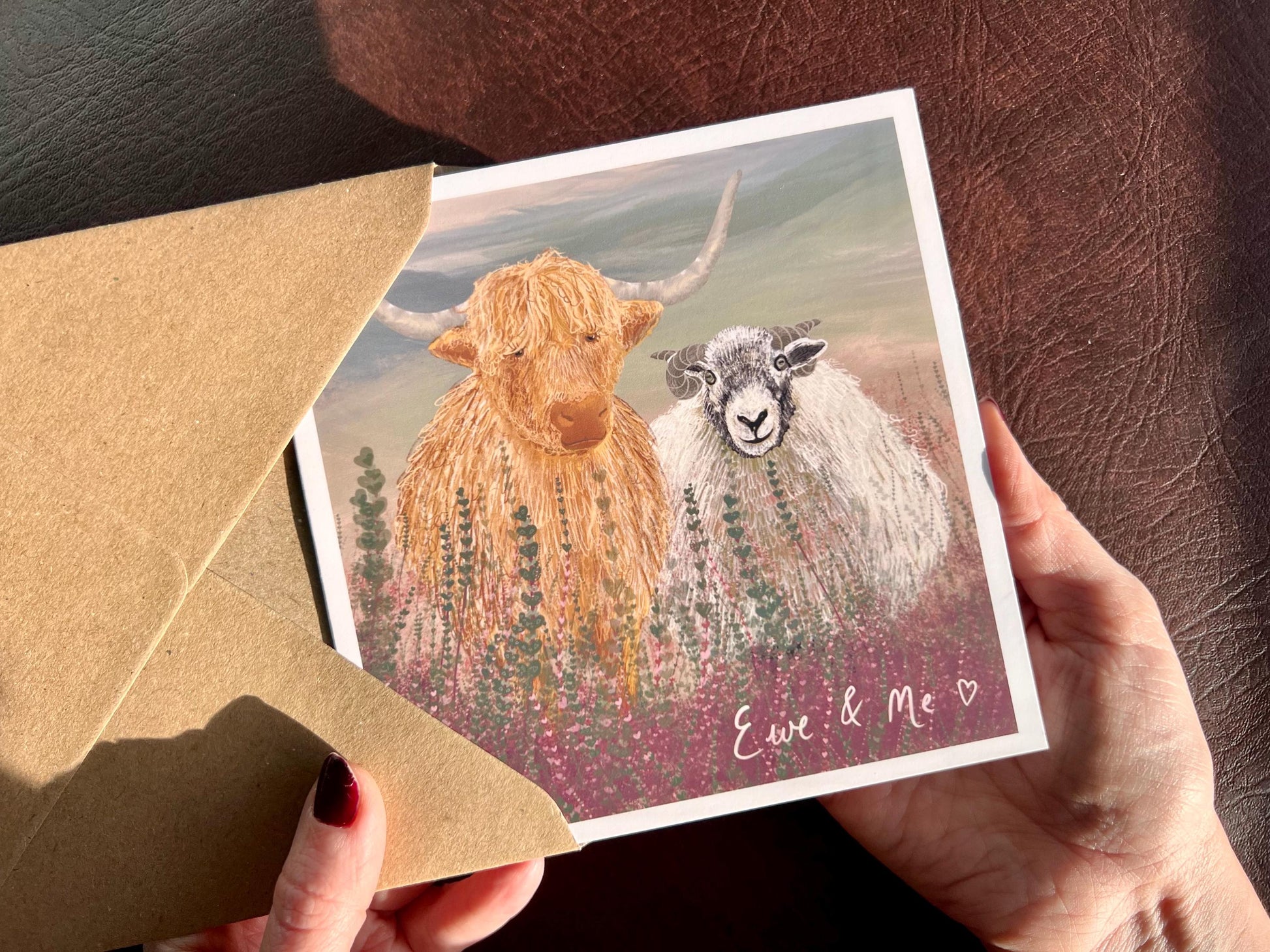 Hands holding an artisan Valentines or anniversary card being removed from a kraft envelope, featuring a playful illustration of a highland cow and Swaledale sheep standing among purple heather with a sage green moorland background. The card includes handwritten style text reading 'Ewe & Me' celebrating love, friendship and companionship.