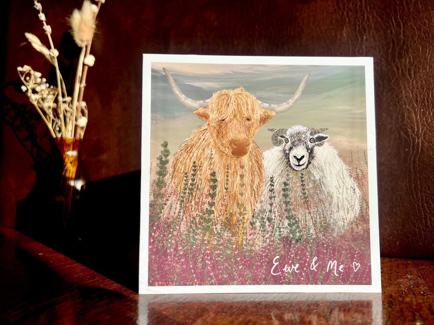 Artisan Valentines or anniversary card featuring a highland cow and Swaledale sheep standing among purple heather with a sage green moorland background and handwritten style text reading 'Ewe & Me', shown standing upright on a wooden tabletop in a styled display.