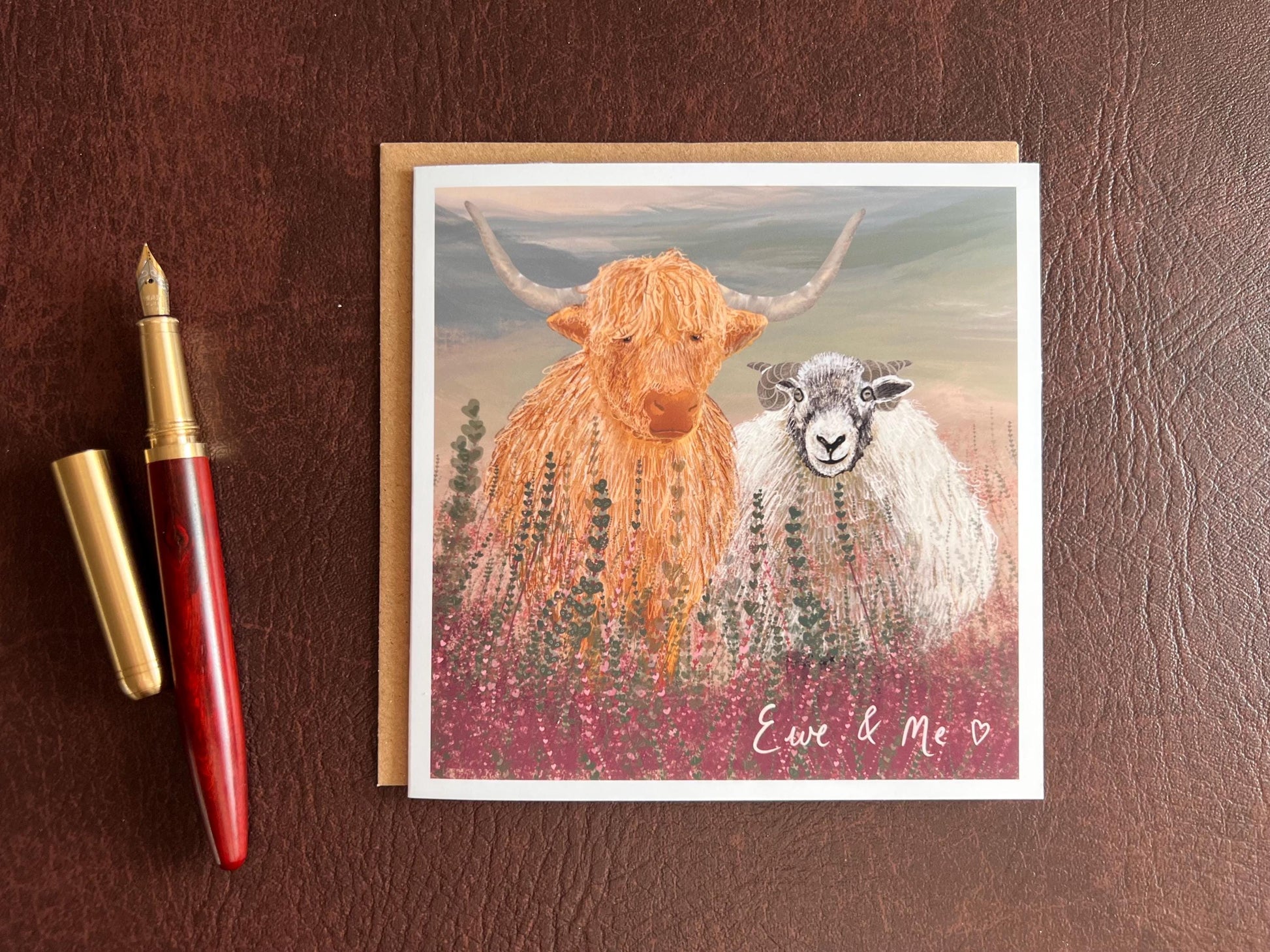 Flat lay image of an artisan Valentines or anniversary card featuring a highland cow and Swaledale sheep standing among purple heather with a sage green moorland background and handwritten style text reading 'Ewe & Me', shown laid flat on a tabletop next to a fountain pen.