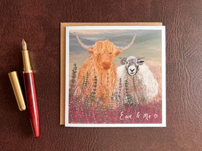 Flat lay image of an artisan Valentines or anniversary card featuring a highland cow and Swaledale sheep standing among purple heather with a sage green moorland background and handwritten style text reading 'Ewe & Me', shown laid flat on a tabletop next to a fountain pen.