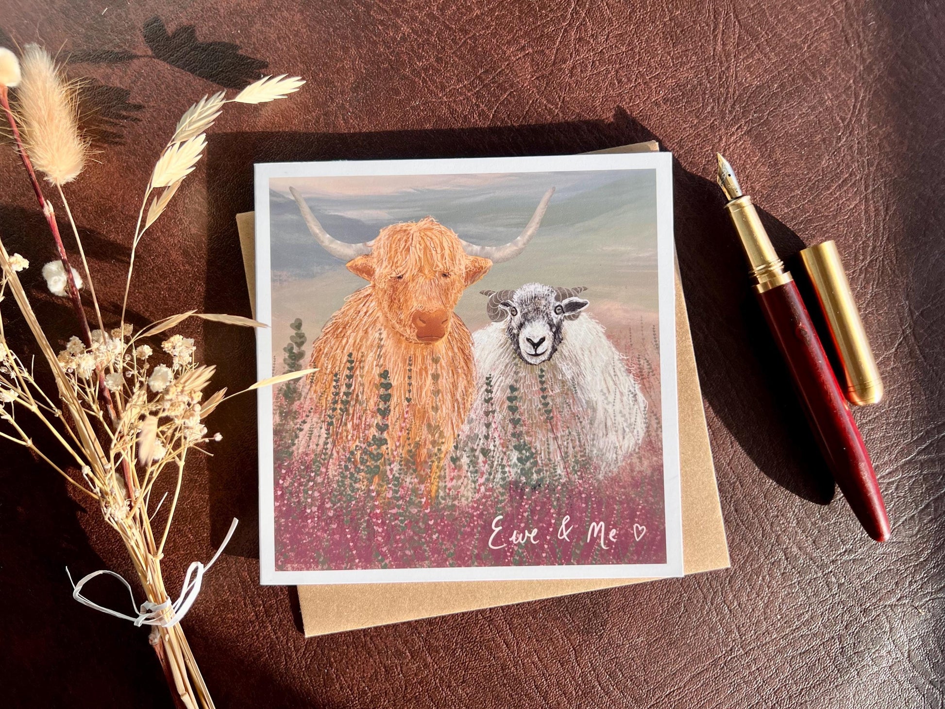 Flat lay image of an artisan Valentines or anniversary card featuring a highland cow and Swaledale sheep standing among purple heather with a sage green moorland background and handwritten style text reading 'Ewe & Me', shown laid flat on a tabletop with a fountain pen and dried flowers in a styled composition.