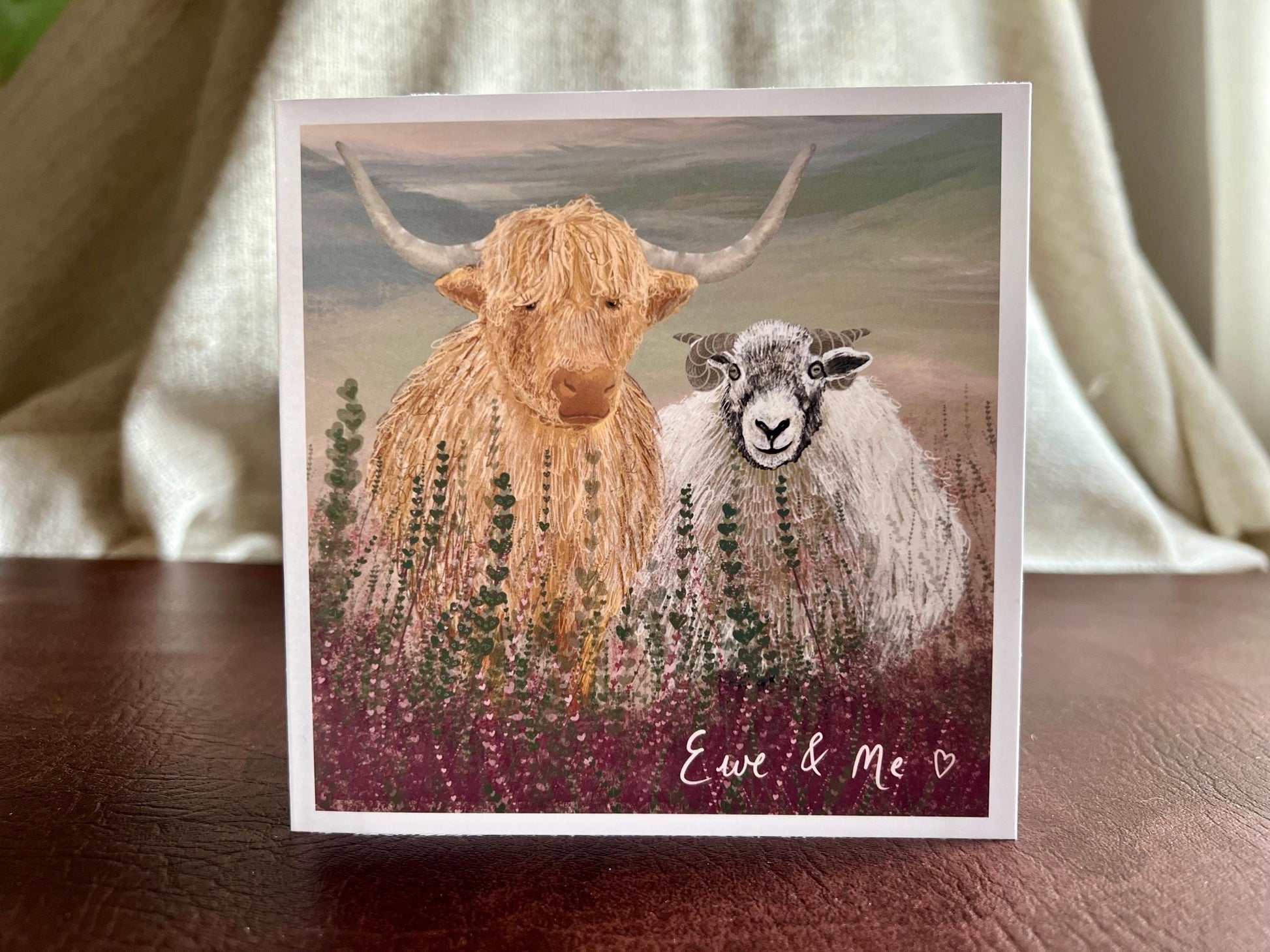Artisan Valentines or anniversary card featuring a highland cow and Swaledale sheep standing among purple heather with a sage green moorland background and handwritten style text reading 'Ewe & Me', shown standing upright on a leatherette tabletop in a styled display.