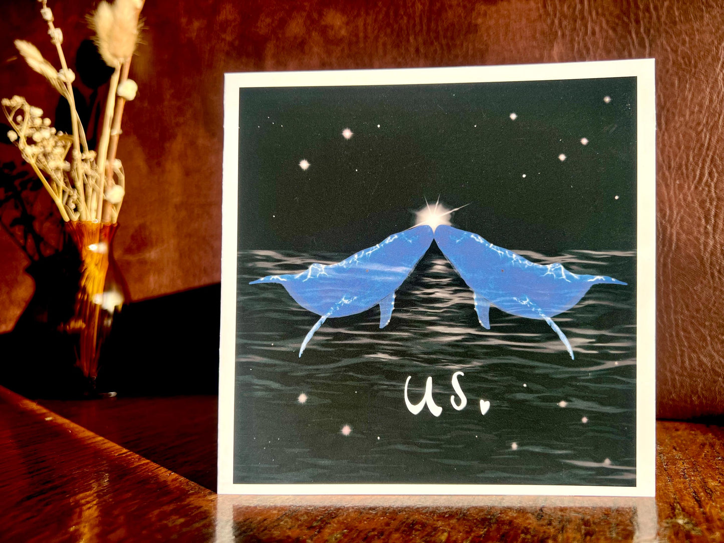 Artisan Valentines or anniversary card featuring two blue whales facing each other nose to nose beneath a starry night sky with a glowing star between them and the word 'us' underneath, shown standing upright on a wooden tabletop in a styled lifestyle setting.