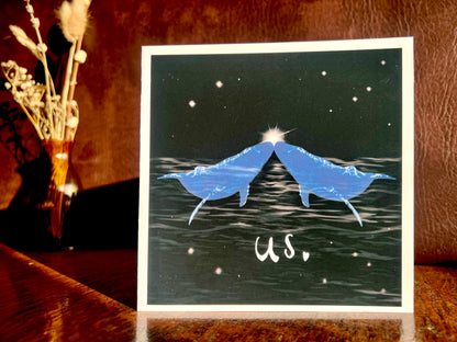 Artisan Valentines or anniversary card featuring two blue whales facing each other nose to nose beneath a starry night sky with a glowing star between them and the word 'us' underneath, shown standing upright on a wooden tabletop in a styled lifestyle setting.