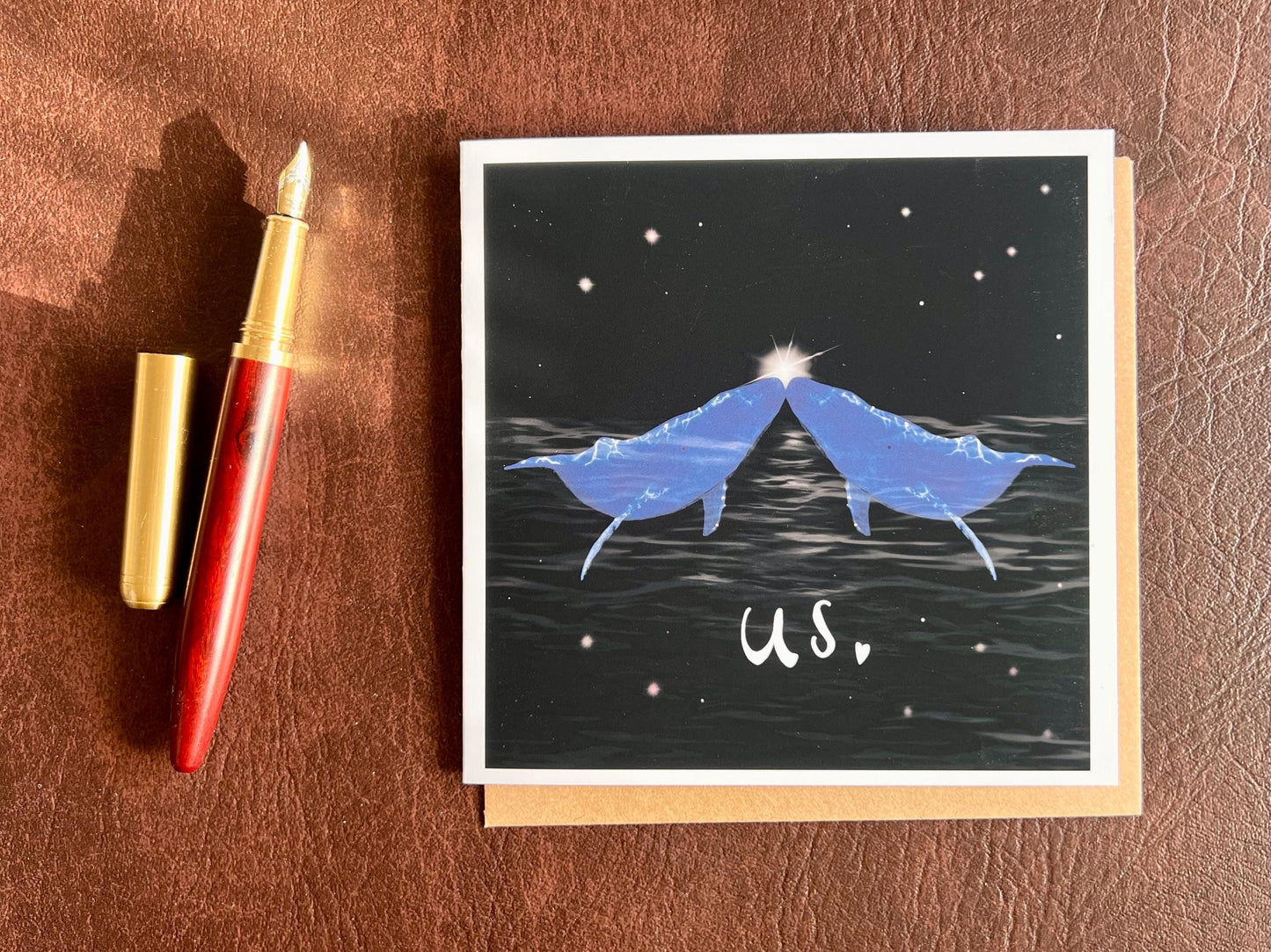 Flat lay image of an artisan Valentines or anniversary card featuring two blue whales facing each other nose to nose beneath a starry night sky with a glowing star between them and the word 'us' underneath, shown laid flat on a tabletop in a styled composition.