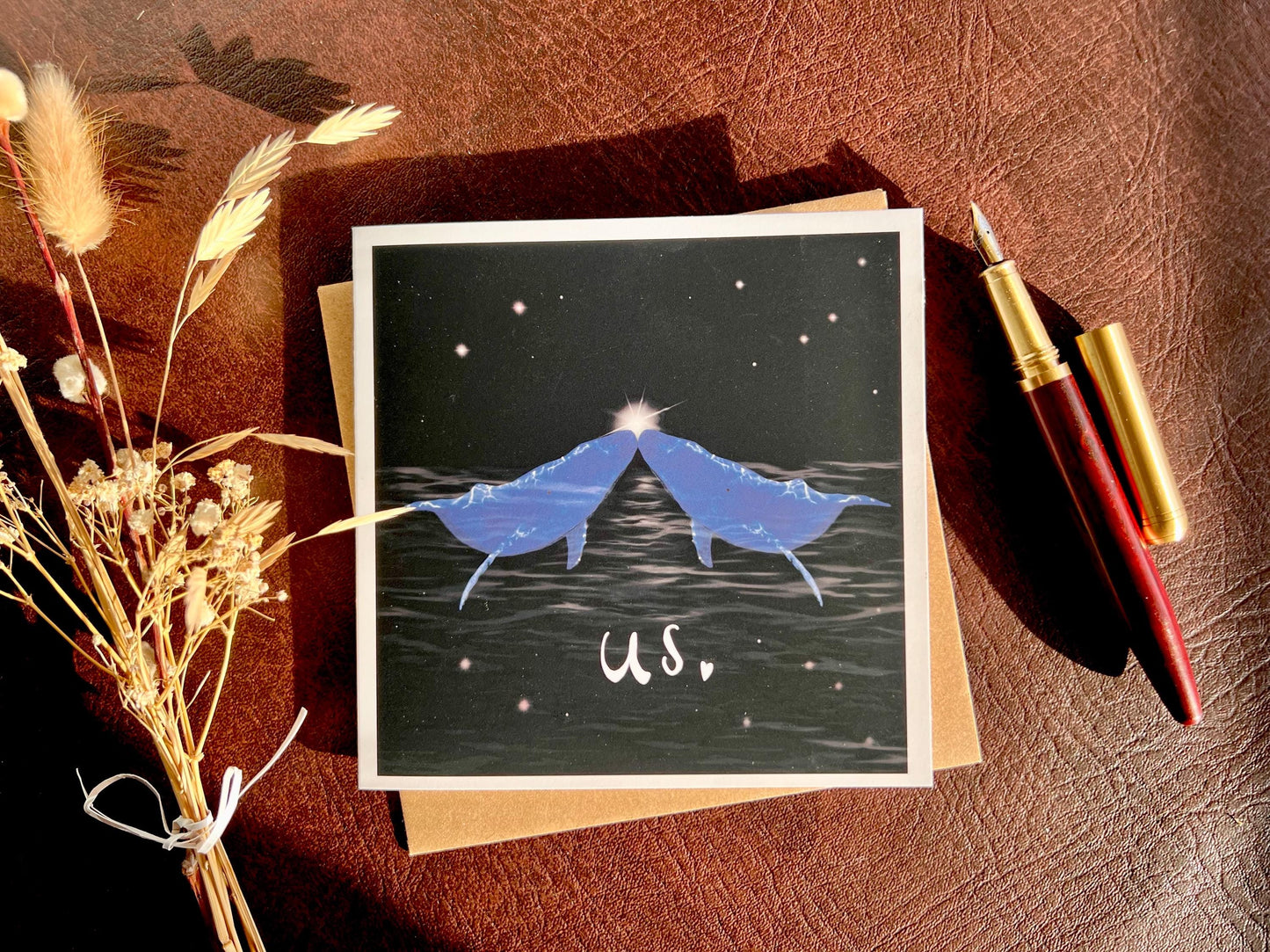 Flat lay image of an artisan Valentines or anniversary card featuring two blue whales facing each other nose to nose beneath a starry night sky with a glowing star between them and the word 'us' underneath, shown laid flat on a tabletop with a fountain pen and dried flowers in a styled composition.