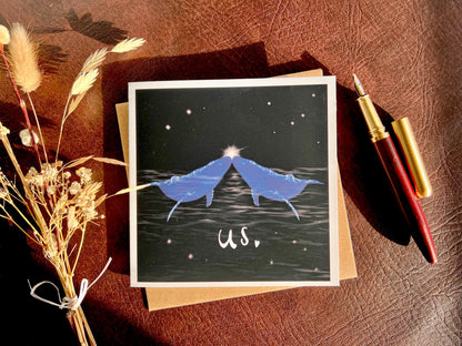 Flat lay image of an artisan Valentines or anniversary card featuring two blue whales facing each other nose to nose beneath a starry night sky with a glowing star between them and the word 'us' underneath, shown laid flat on a tabletop with a fountain pen and dried flowers in a styled composition.