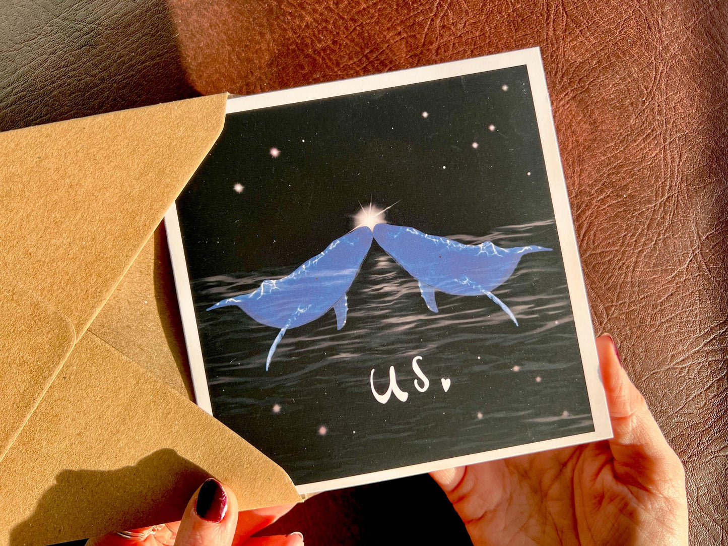 Hands holding an artisan Valentines or anniversary card being removed from a kraft envelope, featuring two blue whales facing each other nose to nose beneath a starry night sky with a glowing star between them. The word 'us' appears underneath in white handwritten style text.