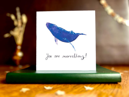 Artisan greeting card featuring a blue whale illustration with a plain white background with handwritten style text 'You are marvellous!', shown standing upright on a green leatherette book in a styled lifestyle display.