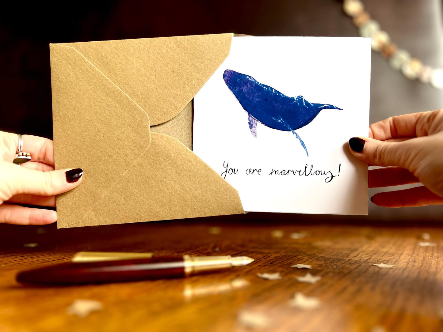 Front view of hands holding an artisan greeting card and kraft envelope, with the card being removed. The design features a blue whale illustration with a plain white background with handwritten style text 'You are marvellous!', created as a positive affirmation card.