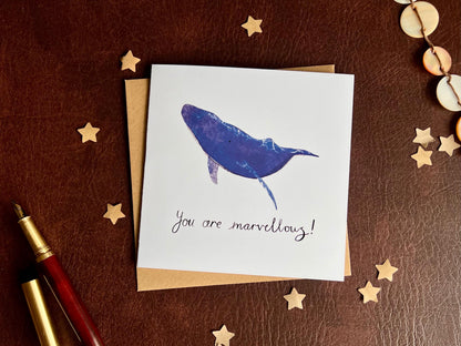 Flat lay staged image of an artisan greeting card featuring a blue whale illustration on a plain white background with handwritten style text 'You are marvellous!', shown laid flat on a leatherette surface next to a pen and surrounded by decorative paper stars.
