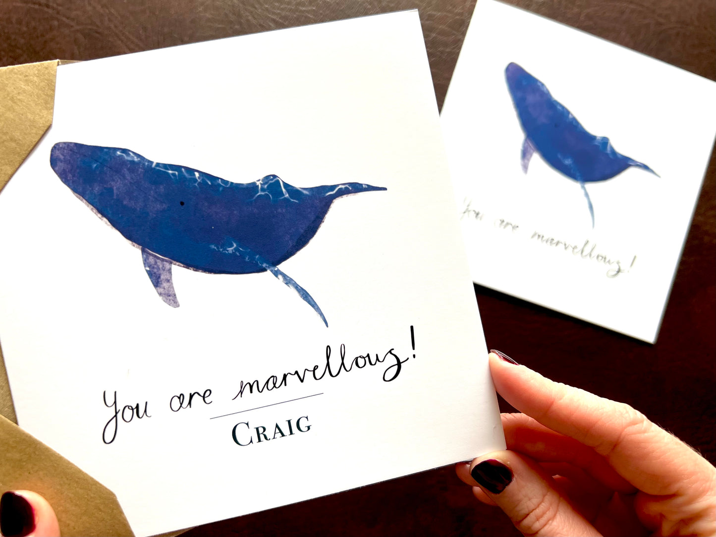 Comparison image showing two artisan whale greeting card designs with a plain white background. The foreground shows the personalised version with a custom name above the handwritten style text 'You are marvellous!', while the background shows the non-personalised version of the same card featuring a blue whale illustration.