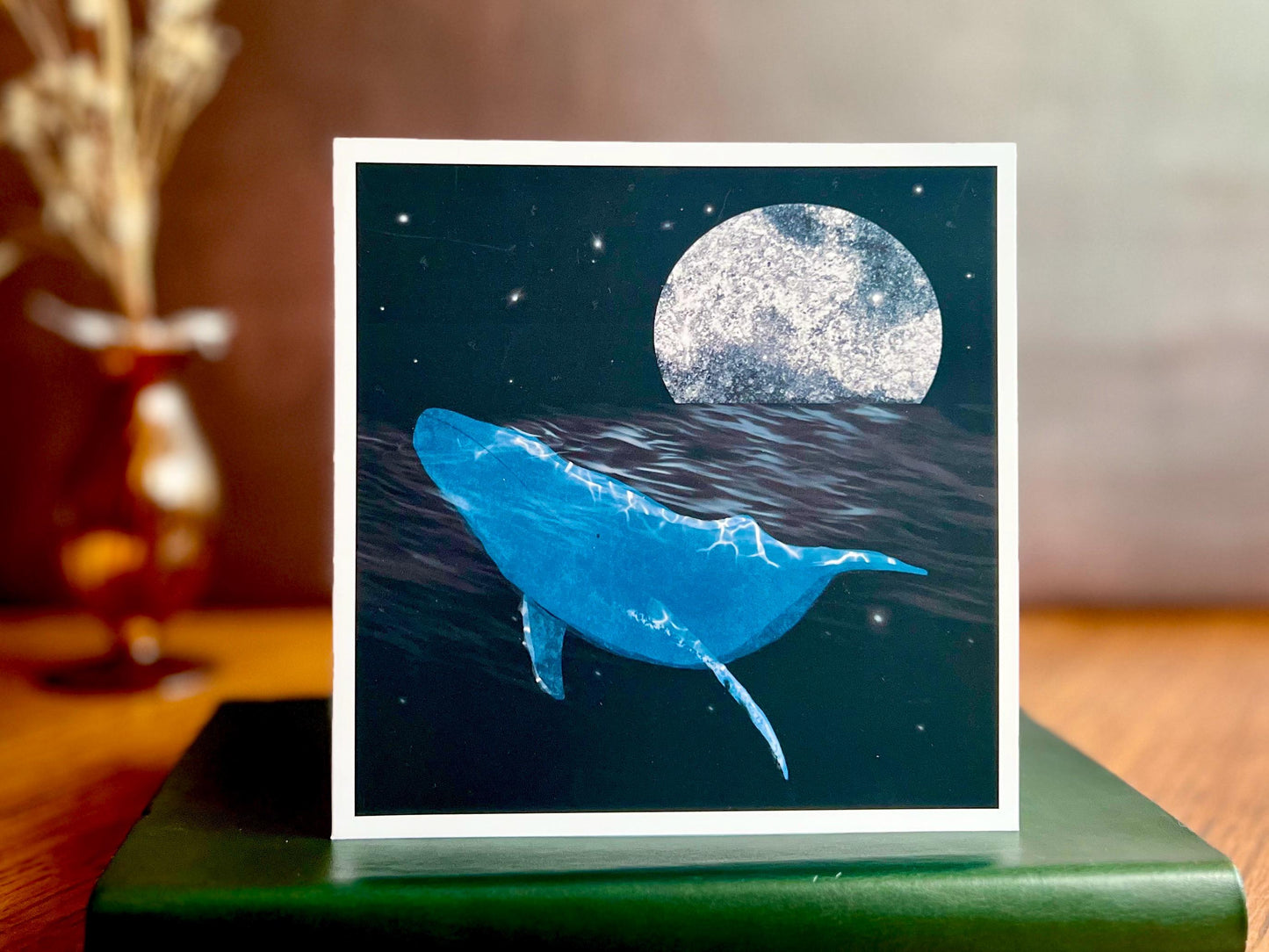 Artisan greeting card featuring a bright blue whale swimming through a dark ocean under a starry night sky with a glowing full moon, displayed standing on a green leatherette book in a styled lifestyle setting.