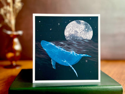Artisan greeting card featuring a bright blue whale swimming through a dark ocean under a starry night sky with a glowing full moon, displayed standing on a green leatherette book in a styled lifestyle setting.