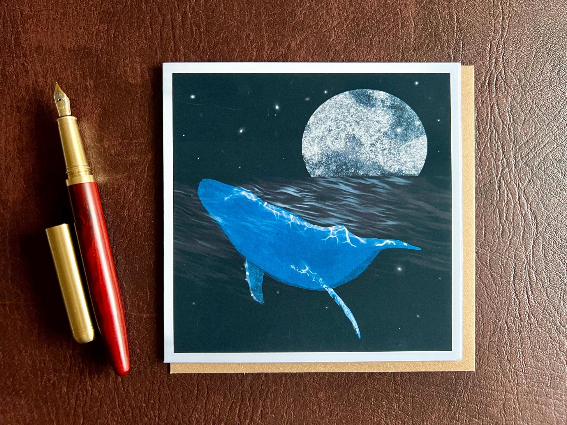 Flat lay image of an artisan greeting card featuring a bright blue whale swimming through a dark ocean under a starry night sky with a glowing full moon, shown laid flat on a tabletop with its kraft envelope in a styled composition.