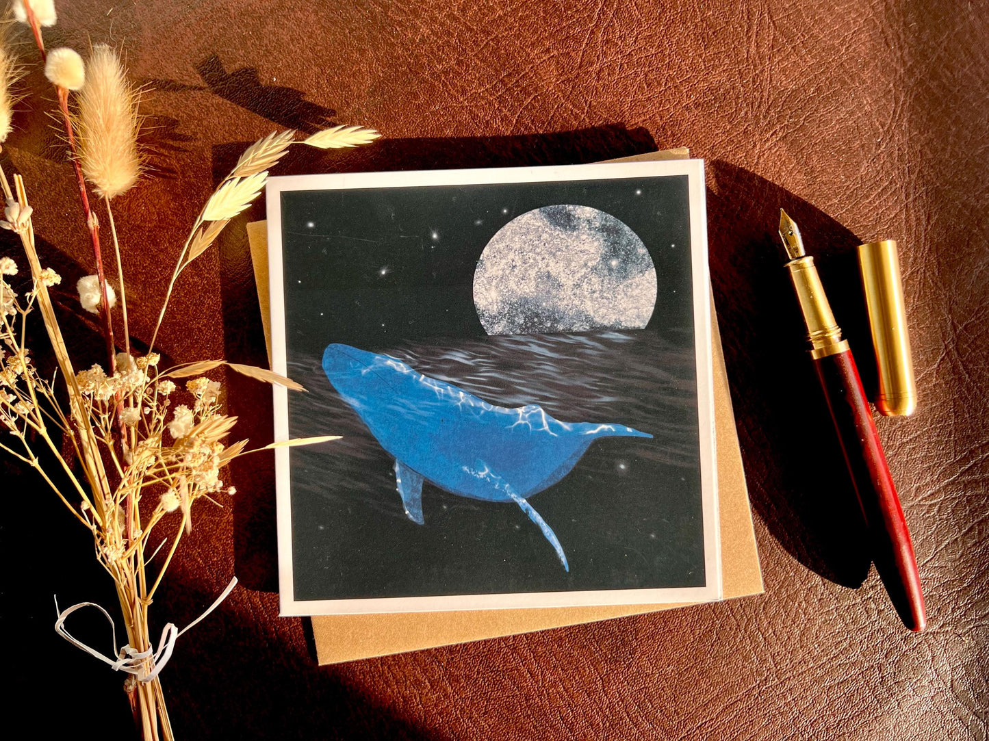 Flat lay image of an artisan greeting card featuring a bright blue whale swimming through a dark ocean under a starry night sky with a glowing full moon, shown laid flat on a tabletop with its kraft envelope, a pen, and dried flowers in a styled composition.