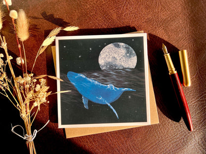 Flat lay image of an artisan greeting card featuring a bright blue whale swimming through a dark ocean under a starry night sky with a glowing full moon, shown laid flat on a tabletop with its kraft envelope, a pen, and dried flowers in a styled composition.