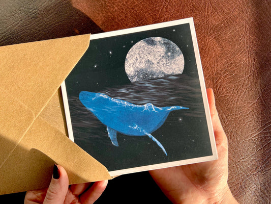 Hands holding an artisan greeting card being removed from a kraft envelope, featuring an illustration of a bright blue whale swimming through a dark ocean beneath a starry night sky with reflections on the water and an ethereal full moon in the background. The card has no text and has a mystical art style suitable for multiple occasions.