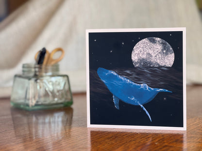 Artisan greeting card featuring a bright blue whale swimming through a dark ocean under a starry night sky with a glowing full moon, displayed upright on a wooden tabletop in a styled lifestyle setting.