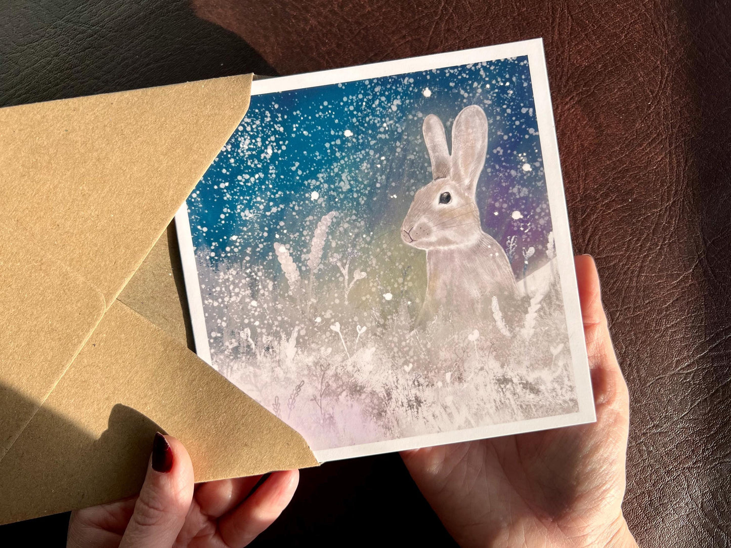 Hands holding an artisan winter card being removed from a kraft envelope, featuring a silvery grey rabbit sitting in snowy foliage beneath a dark blue, green and purple night sky with falling snow and stars. The design has an ethereal Christmas feel.
