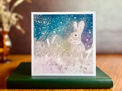 Artisan winter card featuring a silvery grey rabbit sitting in snowy foliage beneath a dark blue, green and purple starry night sky with falling snow, displayed standing on a green leatherette book in a styled lifestyle setting.