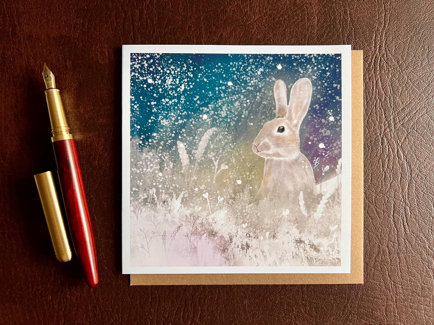 Flat lay image of an artisan winter card featuring a silvery grey rabbit sitting in snowy foliage beneath a dark blue, green and purple starry night sky with falling snow, shown laid flat on a tabletop next to a pen in a styled composition.
