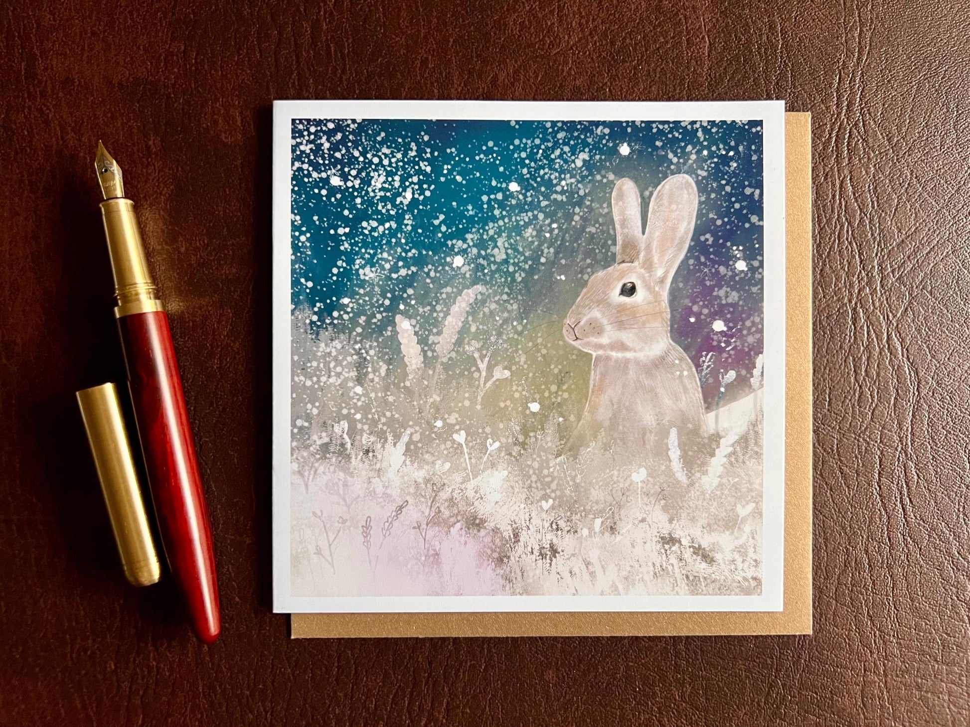Flat lay image of an artisan winter card featuring a silvery grey rabbit sitting in snowy foliage beneath a dark blue, green and purple starry night sky with falling snow, shown laid flat on a tabletop next to a pen in a styled composition.