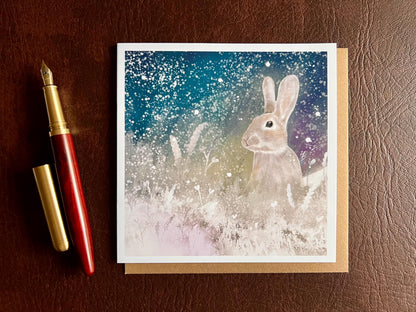 Flat lay image of an artisan winter card featuring a silvery grey rabbit sitting in snowy foliage beneath a dark blue, green and purple starry night sky with falling snow, shown laid flat on a tabletop next to a pen in a styled composition.
