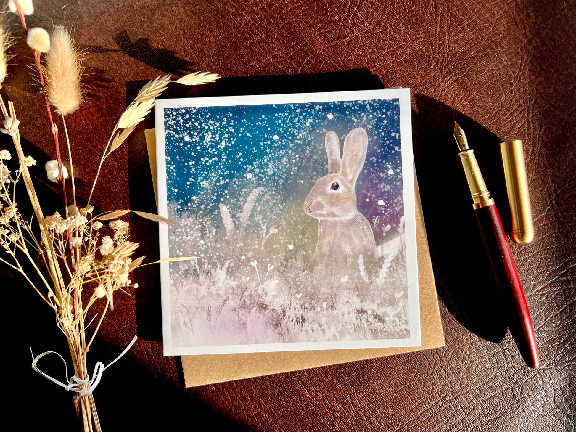 Flat lay image of an artisan winter card featuring a silvery grey rabbit sitting in snowy foliage beneath a dark blue, green and purple starry night sky with falling snow, shown laid flat on a tabletop with a pen and dried flowers in a styled composition.