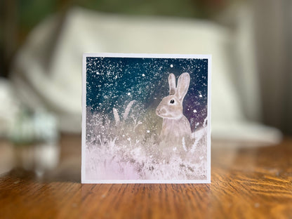 Artisan winter card featuring a silvery grey rabbit sitting in snowy foliage beneath a dark blue, green and purple starry night sky with falling snow, displayed upright on a tabletop with a softly blurred background.