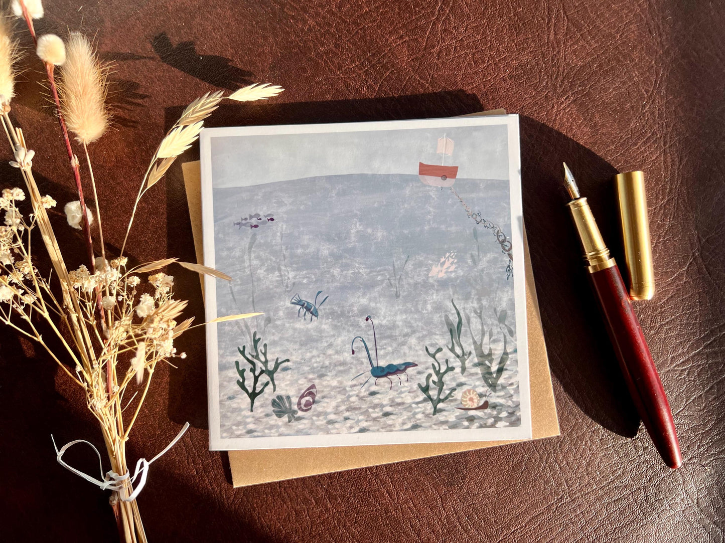 Special illustrated greeting card featuring a blue lobster and blue prawn in a crystal blue underwater coastal scene with a small boat above the water, laid flat and styled with a fountain pen and dried flowers. Joyful summery coastal art greeting card suitable for birthdays or keeping in touch.