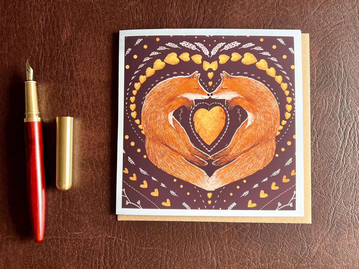 Special illustrated greeting card featuring two foxes sleeping nose-to-nose with their bodies curled into a heart shape, surrounded by a symmetrical leaf and heart pattern on a deep maroon red background. Romantic love-themed card suitable for Valentine’s Day or anniversaries, laid flat for display.