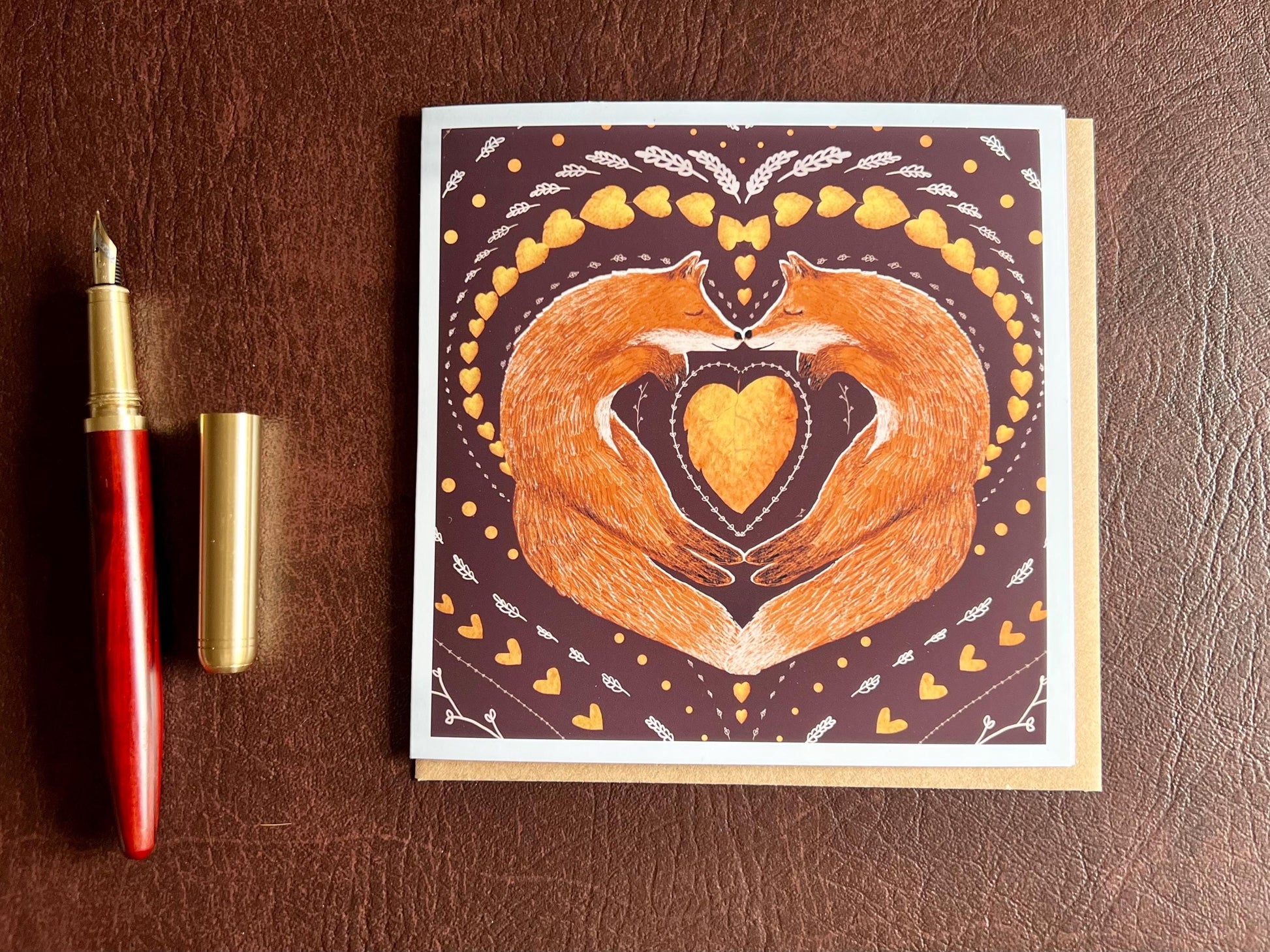 Special illustrated greeting card featuring two foxes sleeping nose-to-nose with their bodies curled into a heart shape, surrounded by a symmetrical leaf and heart pattern on a deep maroon red background. Romantic love-themed card suitable for Valentine’s Day or anniversaries, laid flat for display.