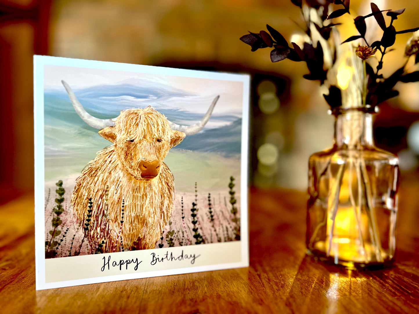 Special illustrated birthday greeting card featuring a highland cow standing amongst heather in a tranquil countryside landscape, displayed upright on a tabletop. The card includes the text “Happy Birthday” beneath the illustration.