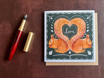 Special illustrated greeting card featuring two red squirrels forming a heart shape with their tails and the word “love” in handwritten font, laid flat on a leatherette tabletop. Romantic Valentine’s Day and anniversary greeting card with dark green background and white scalloped border design.